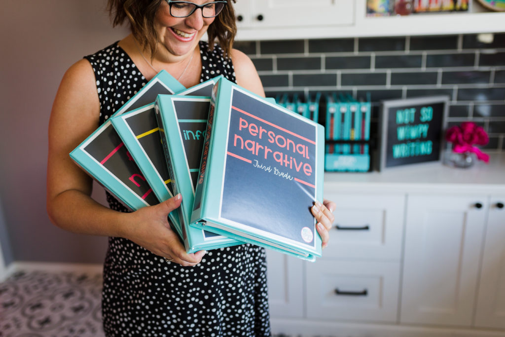 teacher holding 4 binders, one for each genre of writing that can help increase writing test scores.